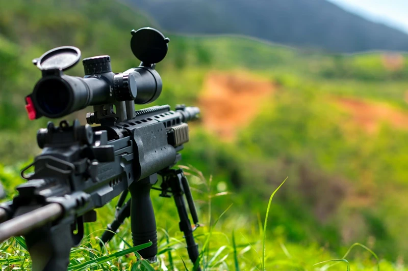 Scoped rifle overlooking grassy hillside trail.